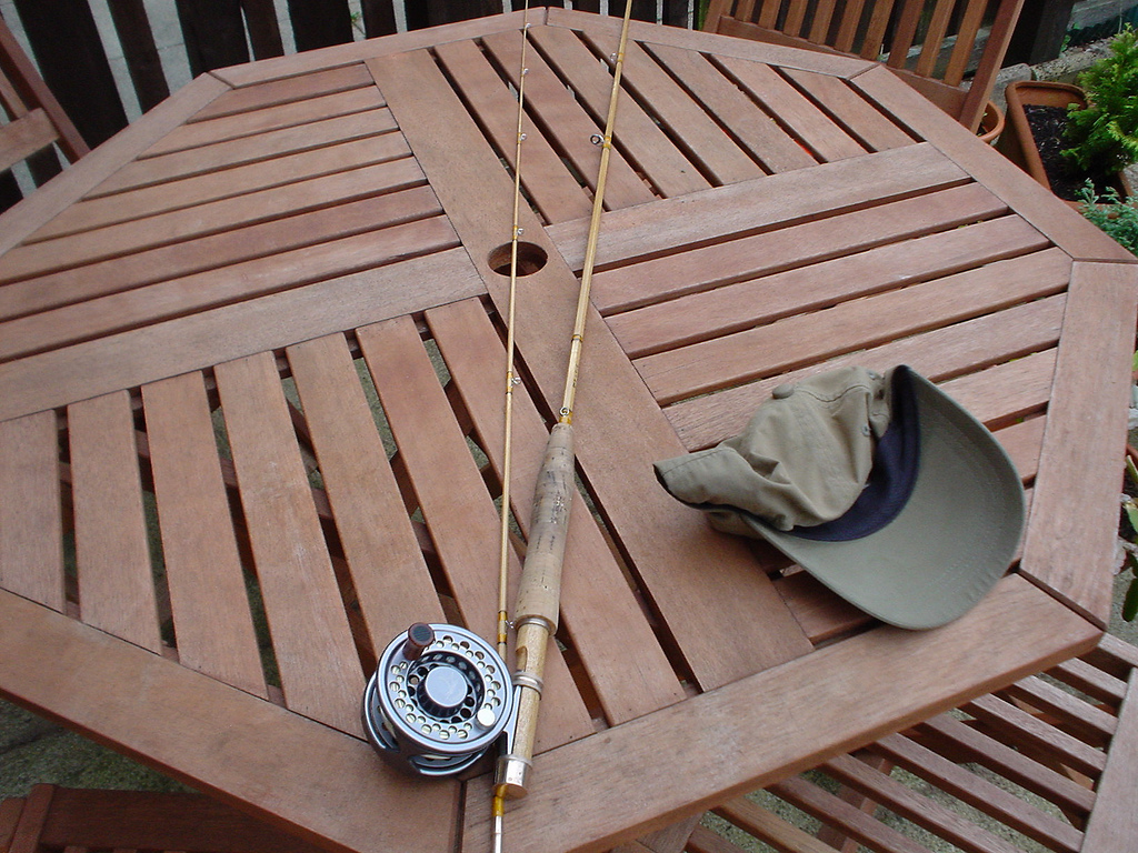 A blonde split-cane fly rod in two sections with a cork grip and a silver large-arbor fly reel, laid across an octagonal slatted wooden garden table next to an olive ball cap