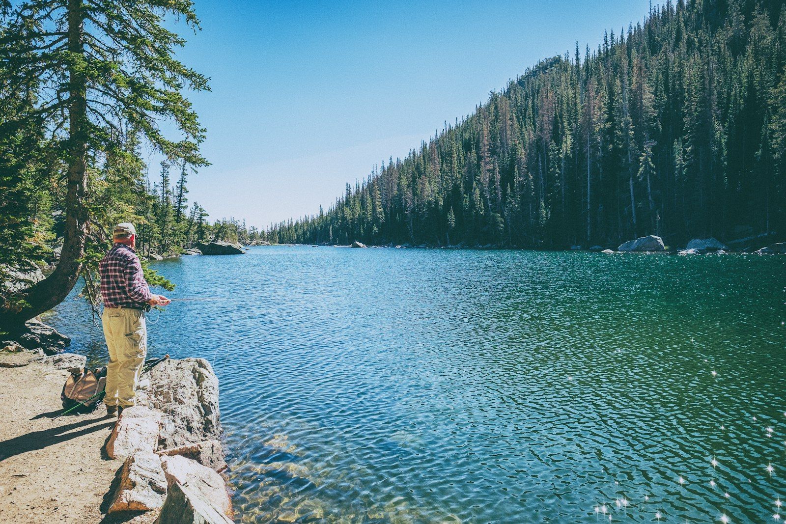 An angler fishing a clear alpine lake ringed in pines — prime brook trout water at the end of a trail