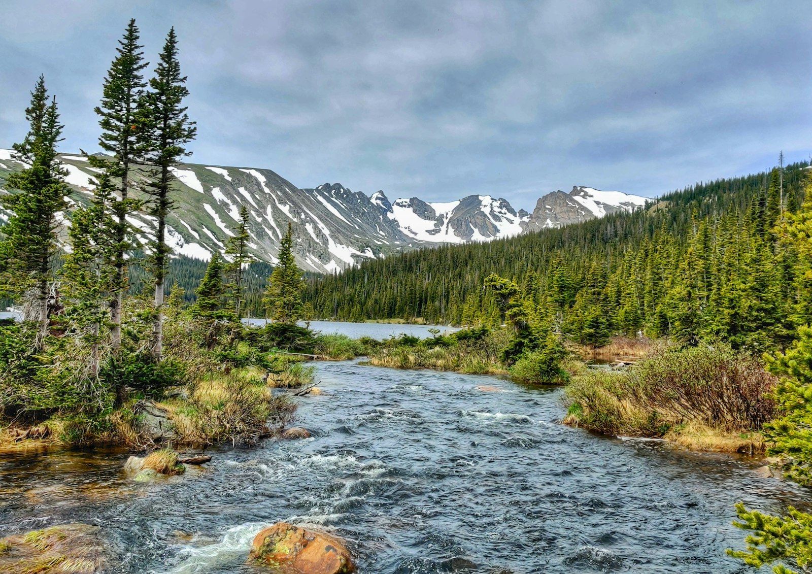 A high-country Colorado creek running out of a lake basin, snow-streaked peaks on the skyline above dark spruce — classic brook trout water