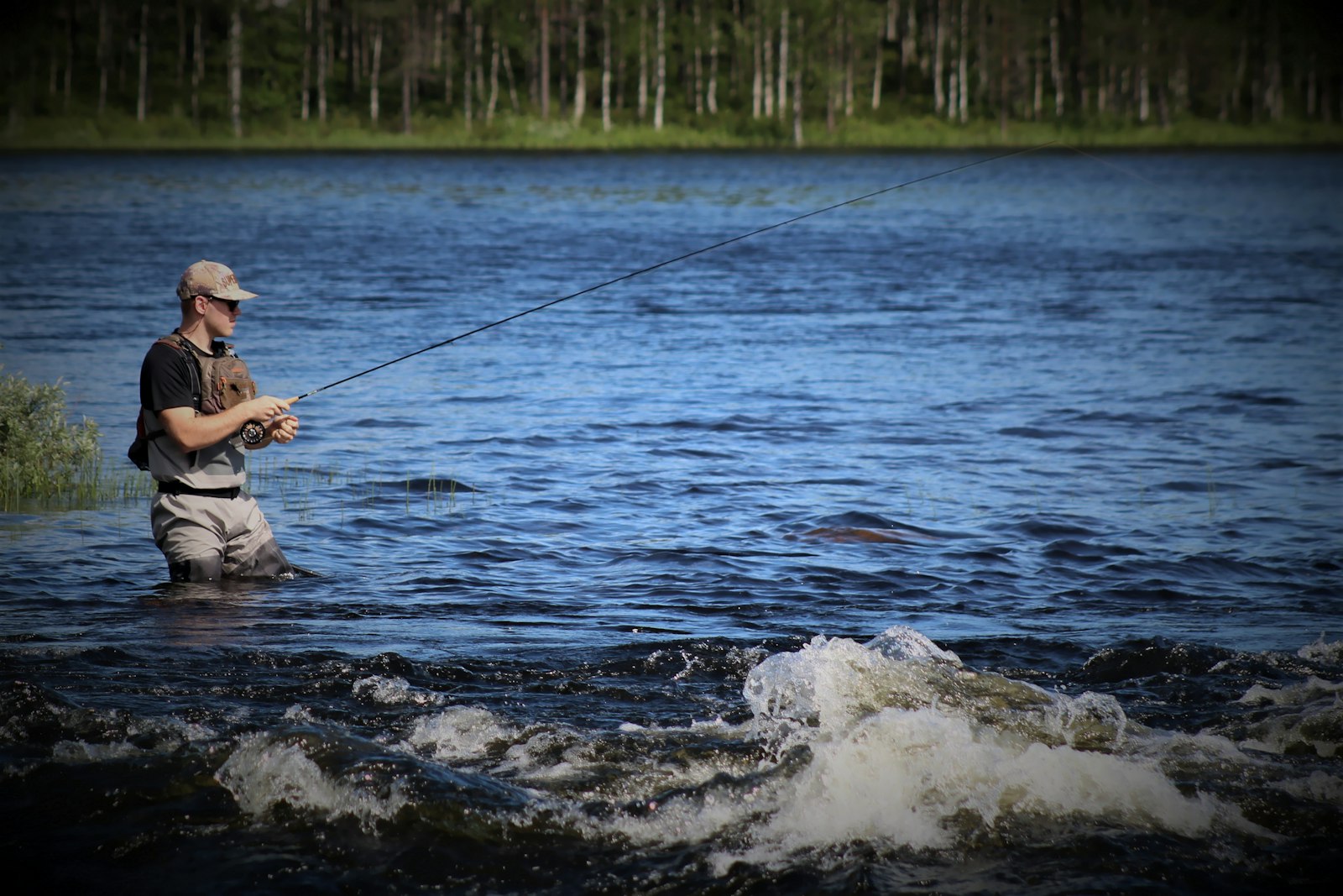 A fly angler standing thigh-deep at the edge of a broad blue river, fly rod held out to one side with line running off the tip, whitewater breaking in the foreground and a birch-and-spruce shoreline behind