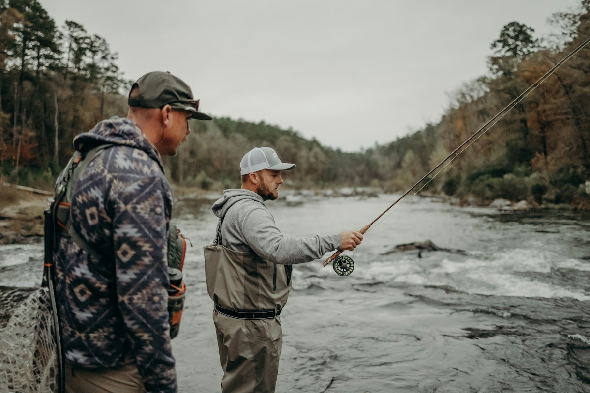 Two anglers in waders at the edge of a broken, rocky river under an overcast sky — one holding a fly rod out over the current with the reel visible, the other standing behind him with a rubber landing net on his back (photo: Unsplash)