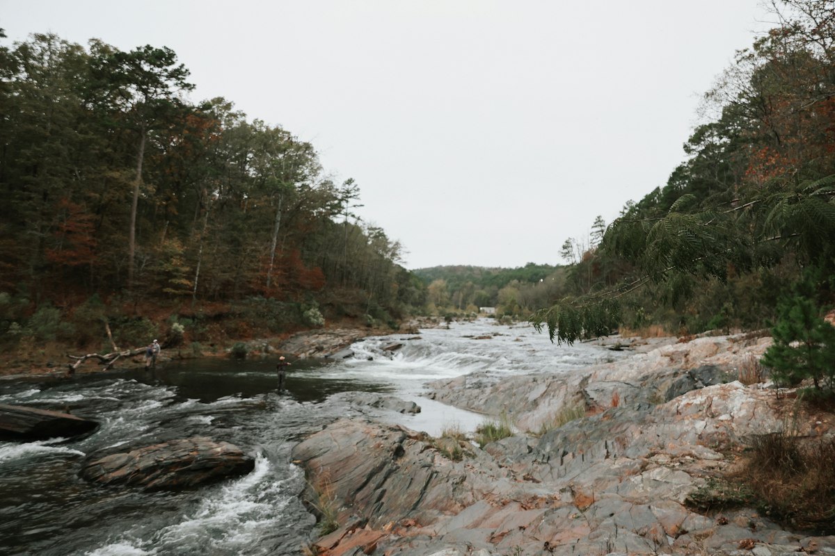 A wide view down a river running over broad bedrock ledges and whitewater chutes, autumn-coloured forest on both banks and two distant anglers standing in the flat water on the left (photo: Unsplash)