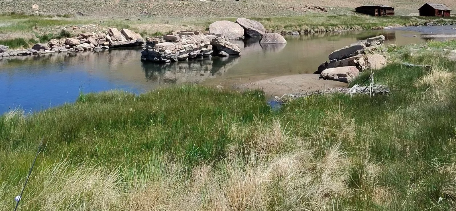 The Dream Stream close up under midday sun — thin, slow water spread wide across an exposed rock bar, meadow grass running right to the edge on both banks, and nothing casting a shadow on any of it