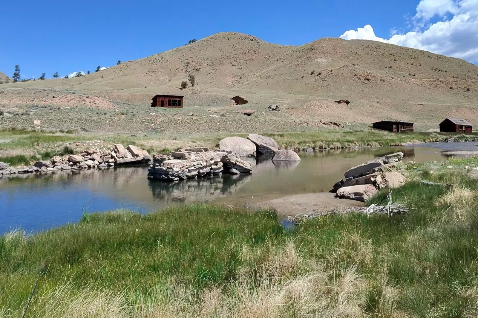 The Dream Stream under a hard blue midday sky — thin, low water spread wide over an exposed rock bar, a bald treeless hill rising straight out of the far bank, old homestead cabins on the slope, and no shade anywhere on the river
