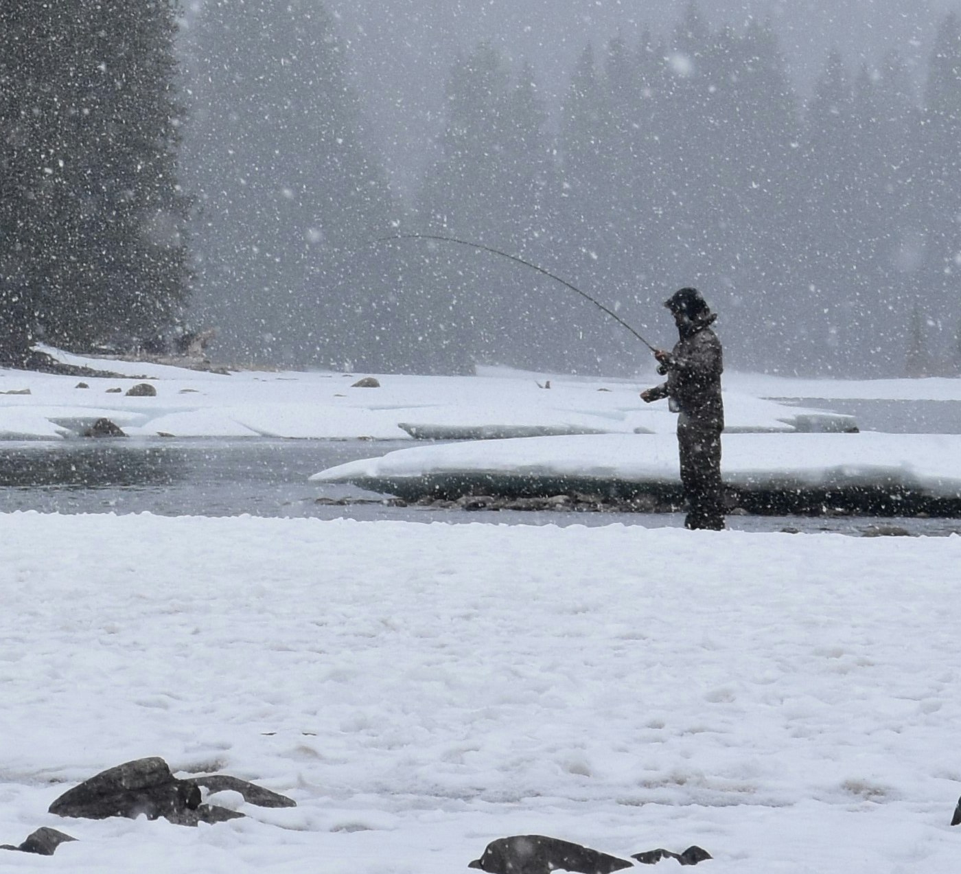 A hooded fly angler stands on a snowy riverbank in heavy falling snow, rod bent in a long arc over dark open water edged with shelf ice (photo: Unsplash)