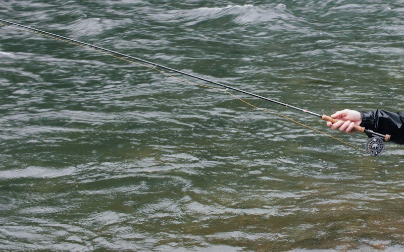 A hand in a wet black rain-jacket sleeve holds a fly rod low over a fast green river, orange-yellow fly line running through the guides to a fly reel below the cork grip (photo: Unsplash)