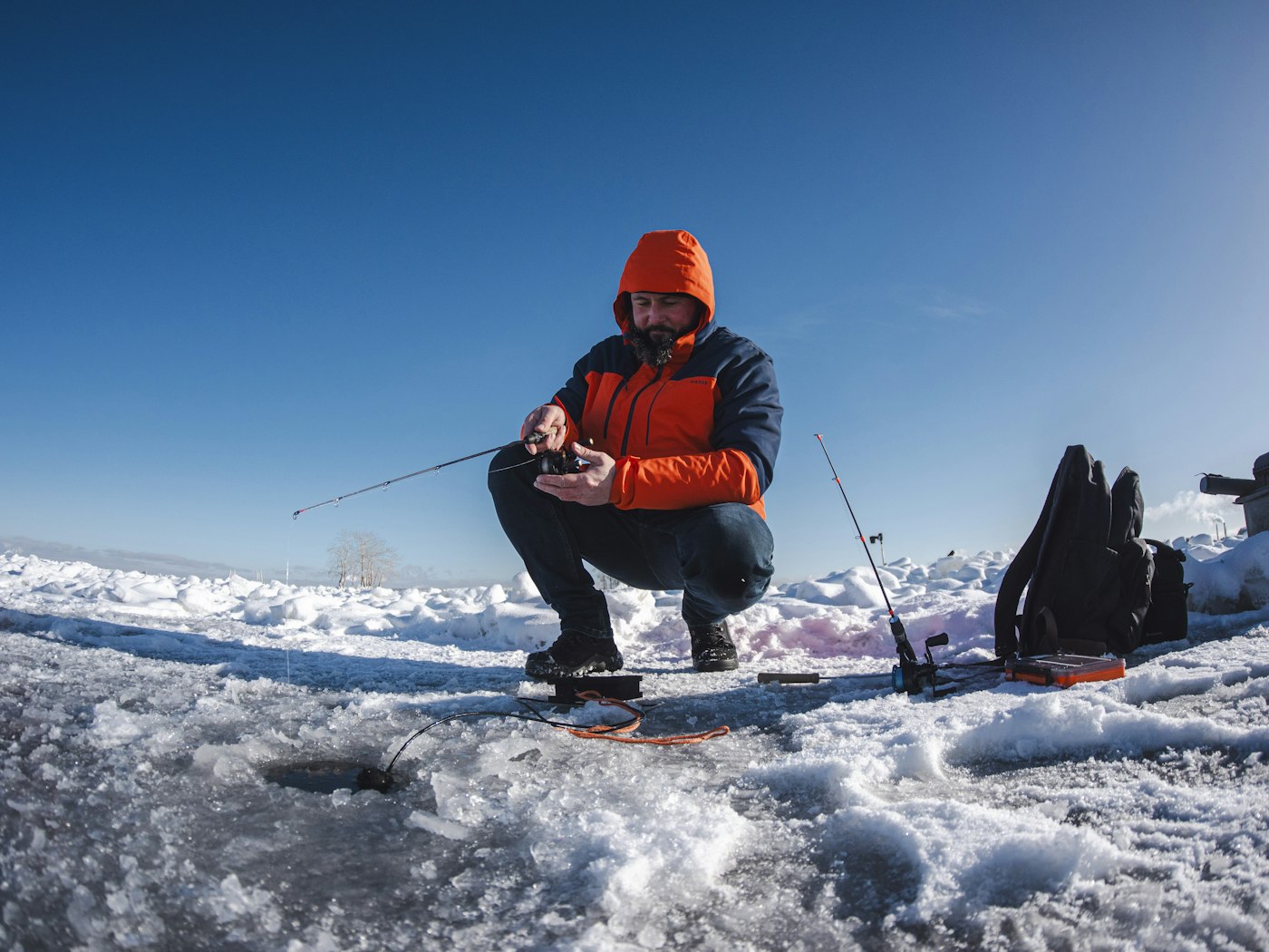 An angler crouched over a fresh hole on snowy ice, a short jigging rod in hand and a sonar transducer cable running down the hole (photo: Unsplash)