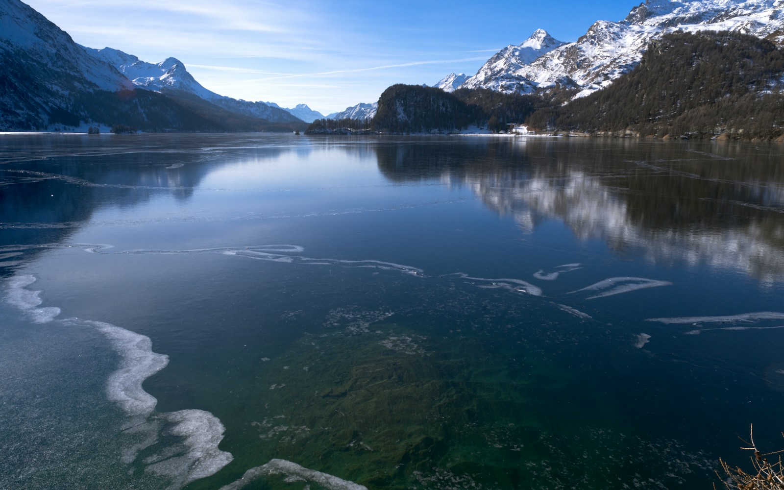A high mountain lake just starting to freeze, thin skim ice spreading across dark open water below snowy peaks