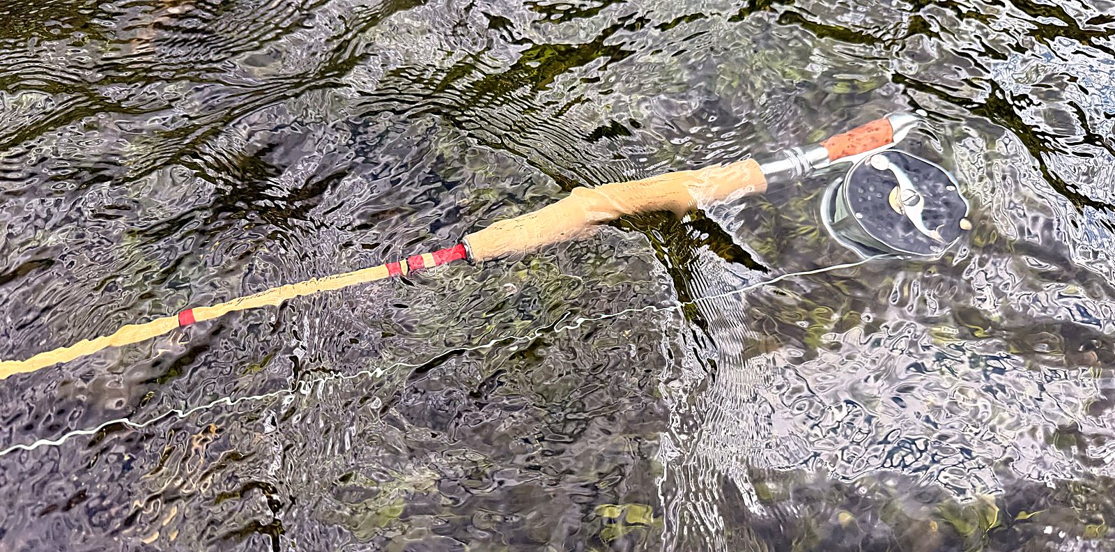 My Gallatin resting in shallow, clear water — blonde cane, red thread wraps, cork grip, downlocking reel seat, and the click-and-pawl reel. The kind of rod that rewards being looked after.