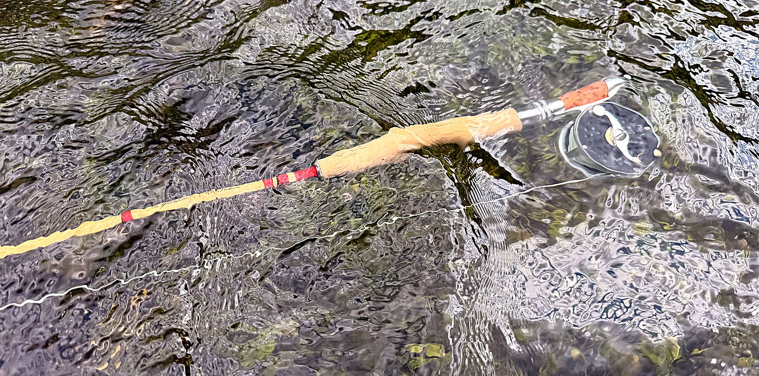 My Gallatin lying in the shallows with the Clark Fork 2.5 mounted — blonde cane, red wraps, cork grip, line trailing in the current