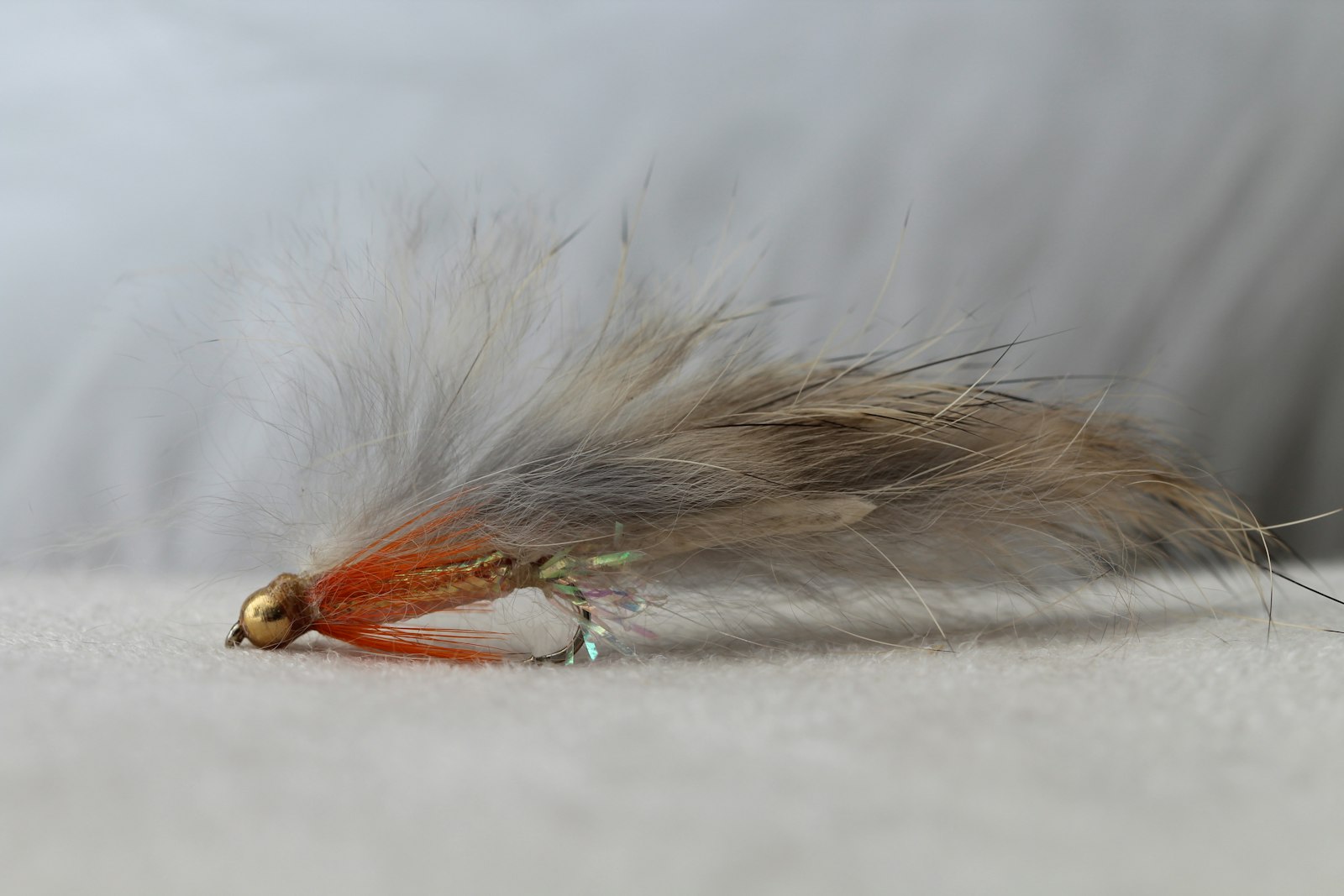 A gold bead-head fly with an orange hot-spot collar, holographic flash and a long soft rabbit-strip wing, laid on pale cloth
