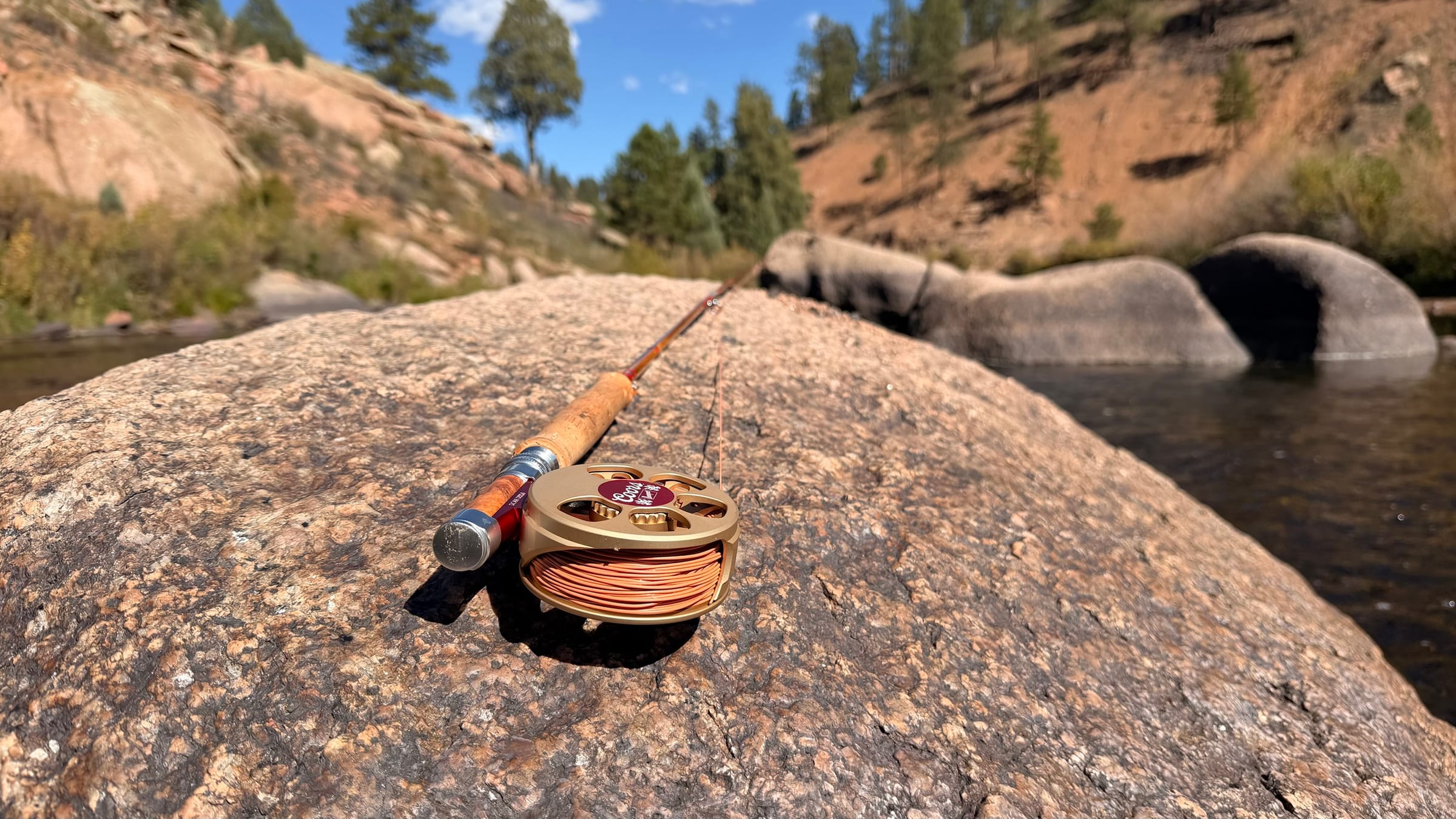 The Ross Coors Banquet Cimarron Creek, buff Coors-gold with its red center circle, mounted on a Headwaters Madison cane rod and resting on a granite boulder beside the river