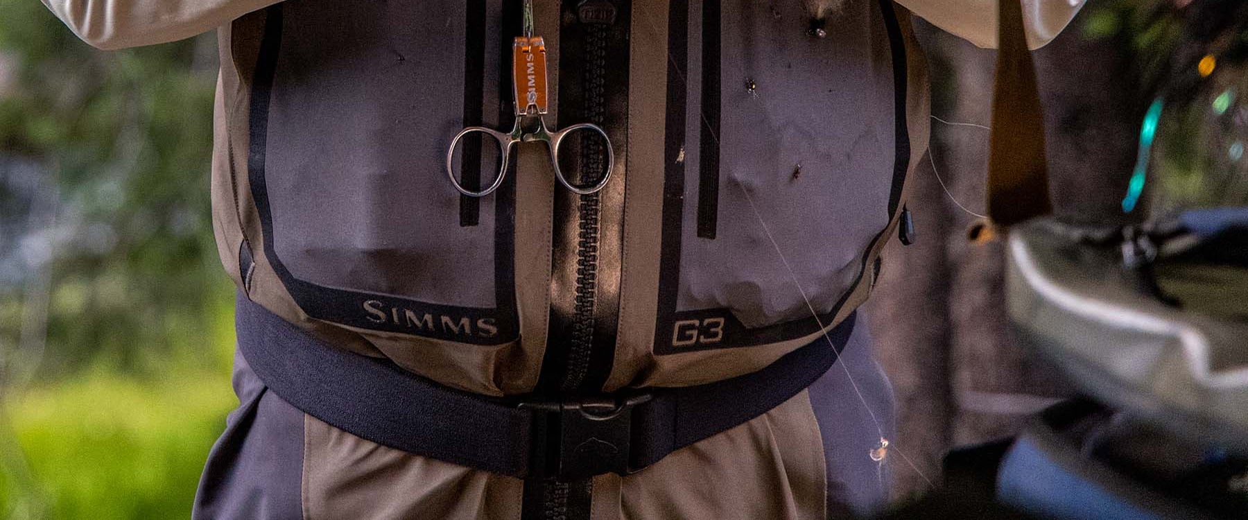 Close-up of a Simms G3Z Pro front-zip wader: the black center zipper, SIMMS and G3 lettering on the chest pockets, forceps on an orange retractor, a few flies stuck to the fabric, and the wading belt (photo: Simms)